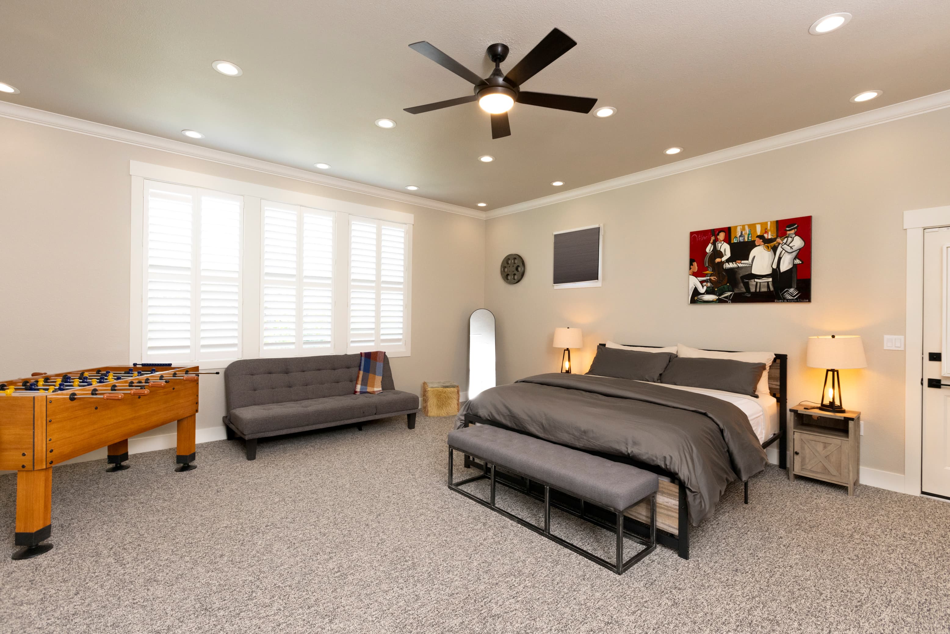 A modern bedroom featuring a bed, seating area, and a foosball table, illuminated by natural light from window shutters.