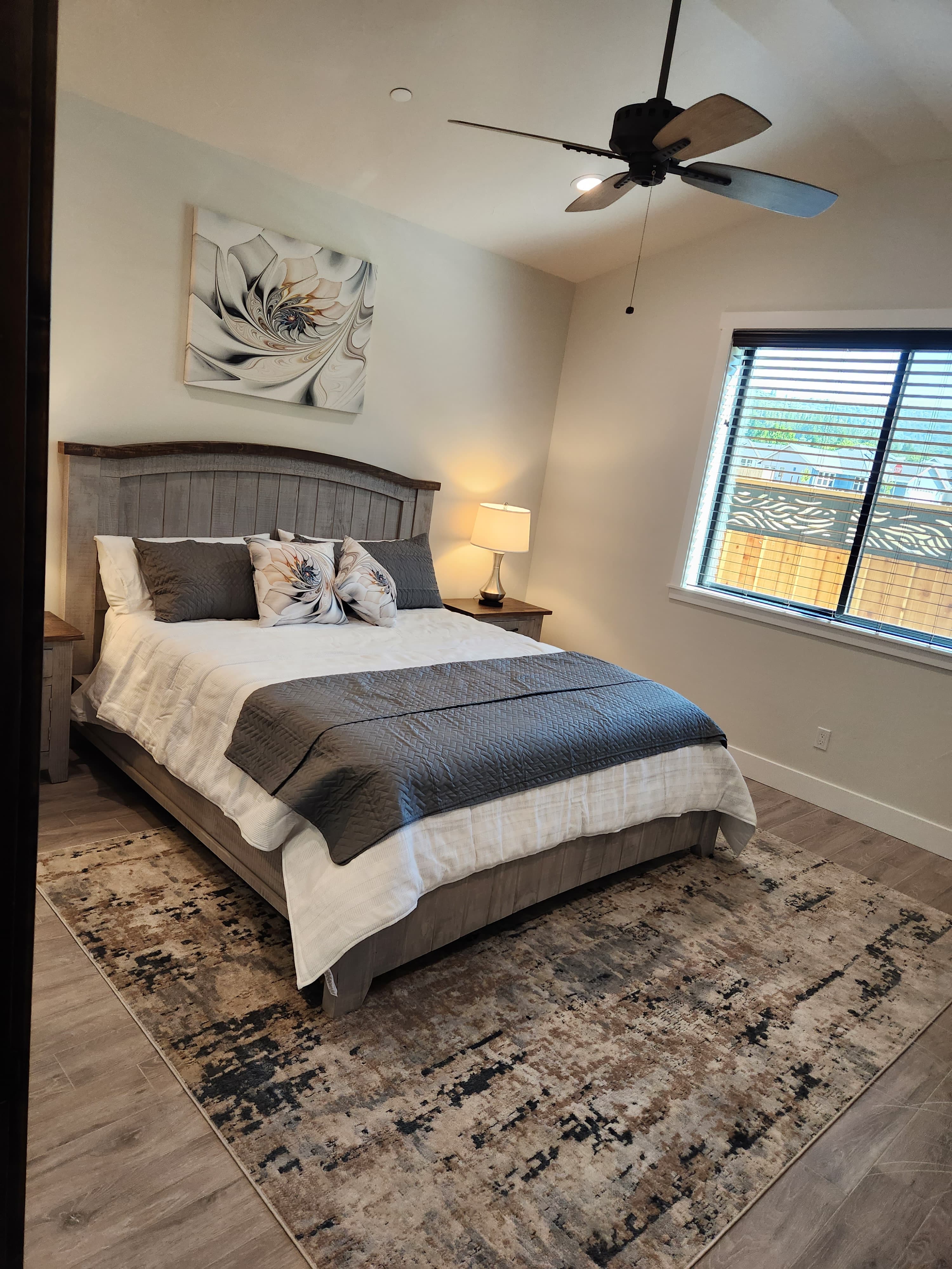 A cozy bedroom featuring a gray bedspread, decorative pillows, a bedside lamp, and a window with blinds.