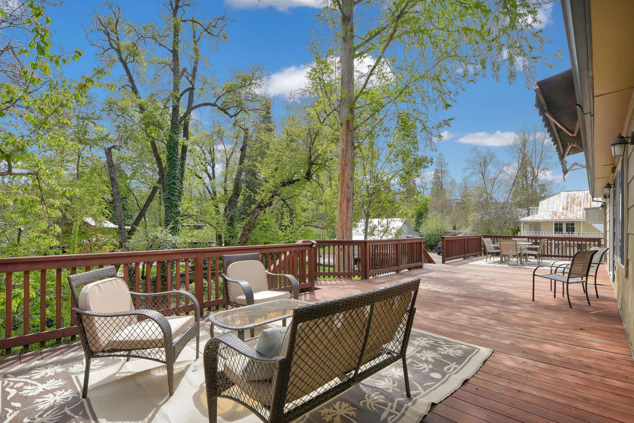 A spacious wooden deck with outdoor seating surrounded by lush greenery under a blue sky.