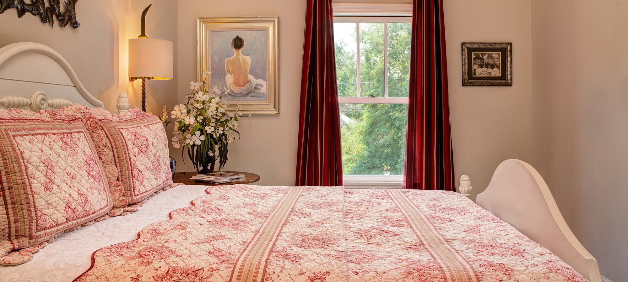 A cozy bedroom featuring a floral-patterned quilt, decorative pillows, and a window with red curtains.