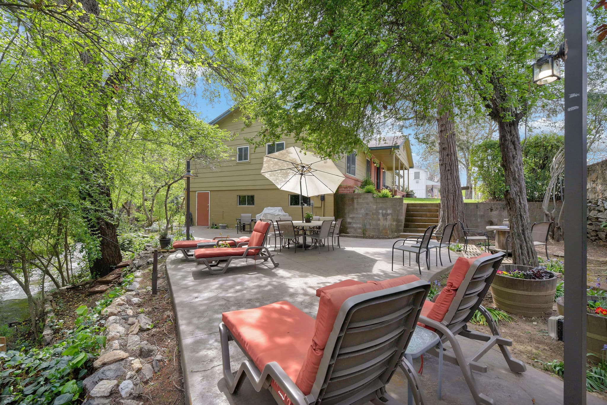 A sunny outdoor patio with lounge chairs, a table, and an umbrella surrounded by greenery and a nearby stream.