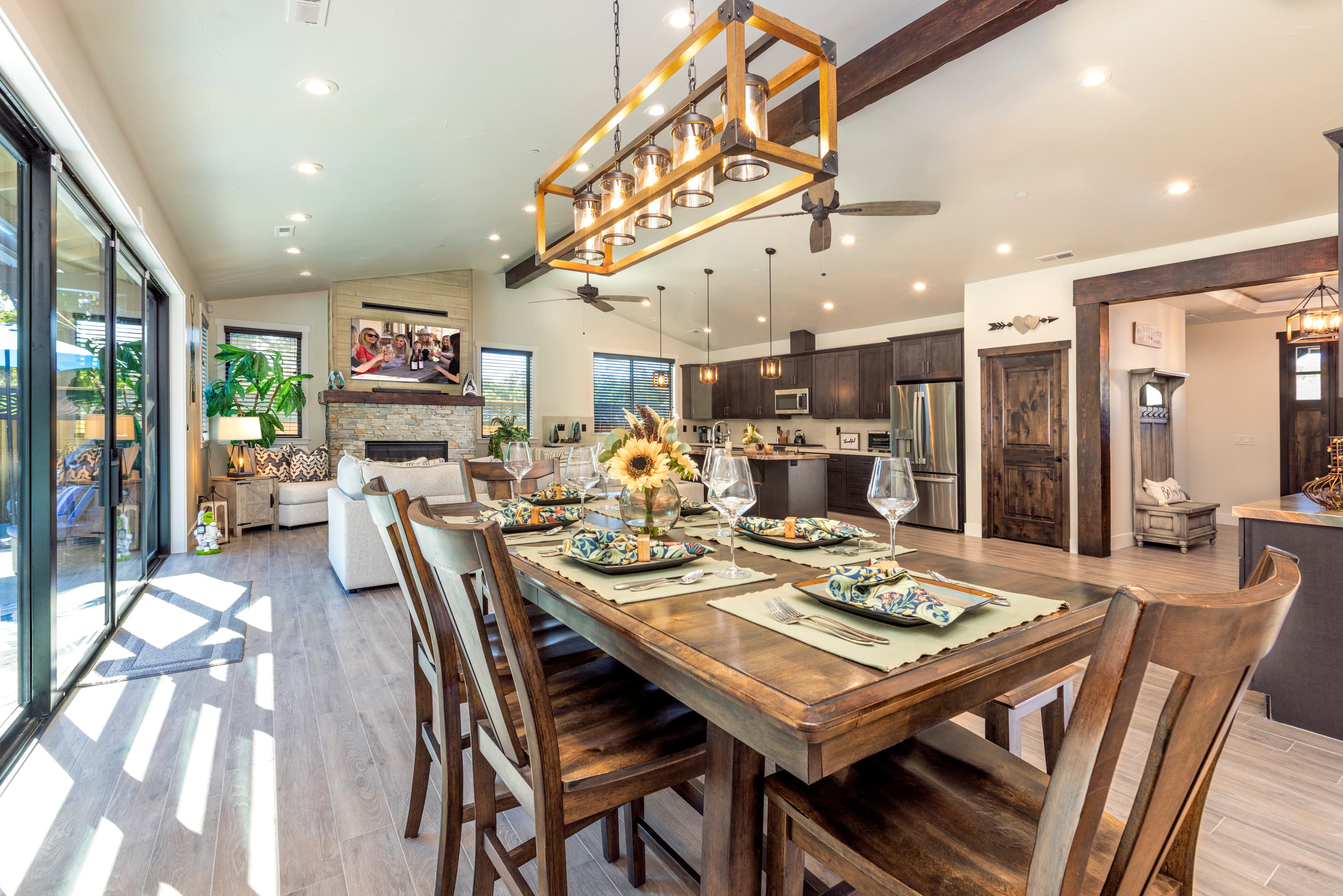 A modern open-concept dining area featuring a wooden table set with dishes, surrounded by stylish chairs and a view of a well-lit kitchen.