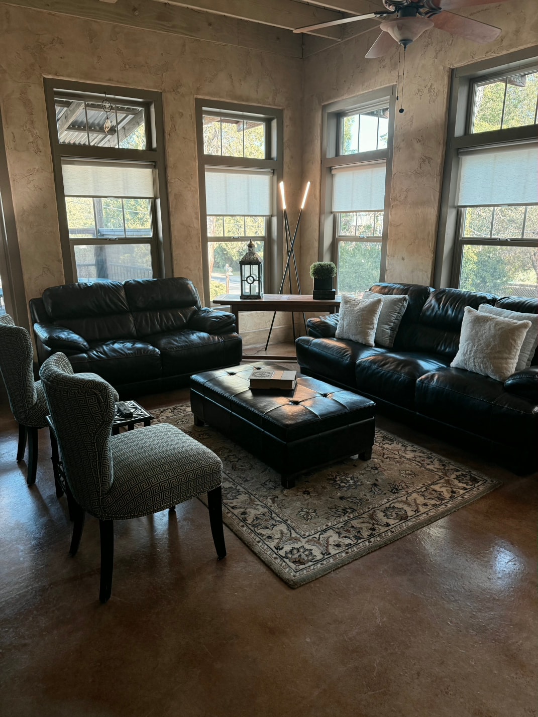 A cozy living room featuring two black leather sofas, a decorative rug, and a coffee table with various items, illuminated by natural light.