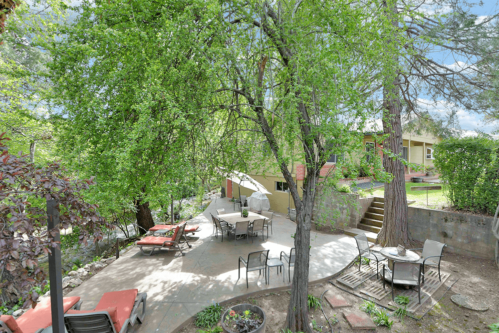 A serene outdoor patio surrounded by lush greenery and a nearby house.