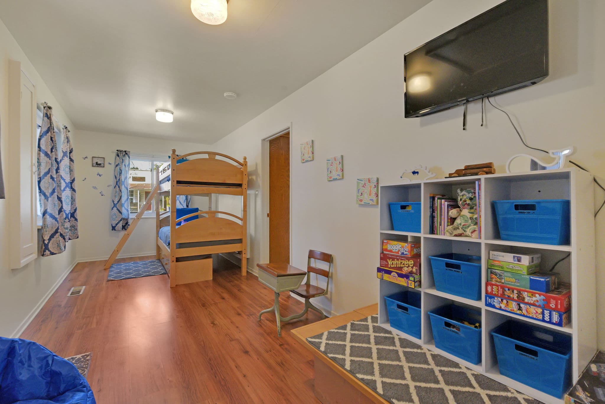 A brightly lit children's bedroom featuring a bunk bed, a wooden chair, storage cubes with toys, and a wall-mounted TV.