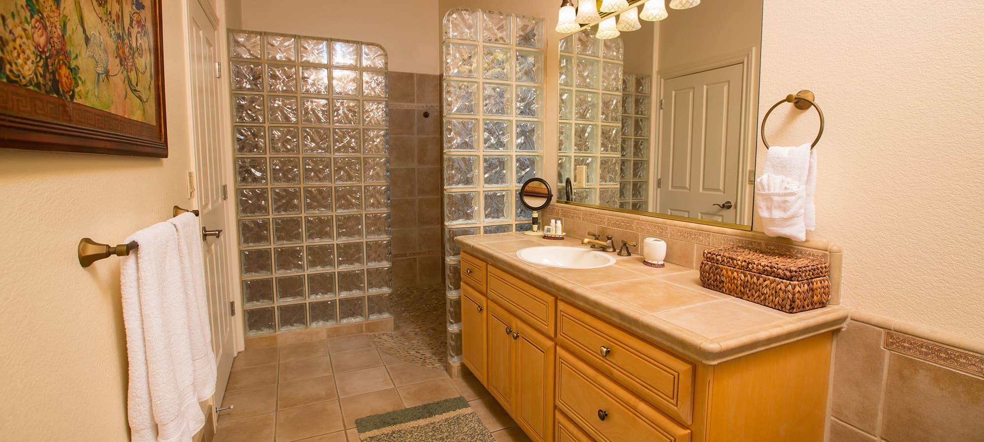 A modern bathroom featuring a double sink vanity, glass block shower, and neutral tile flooring.