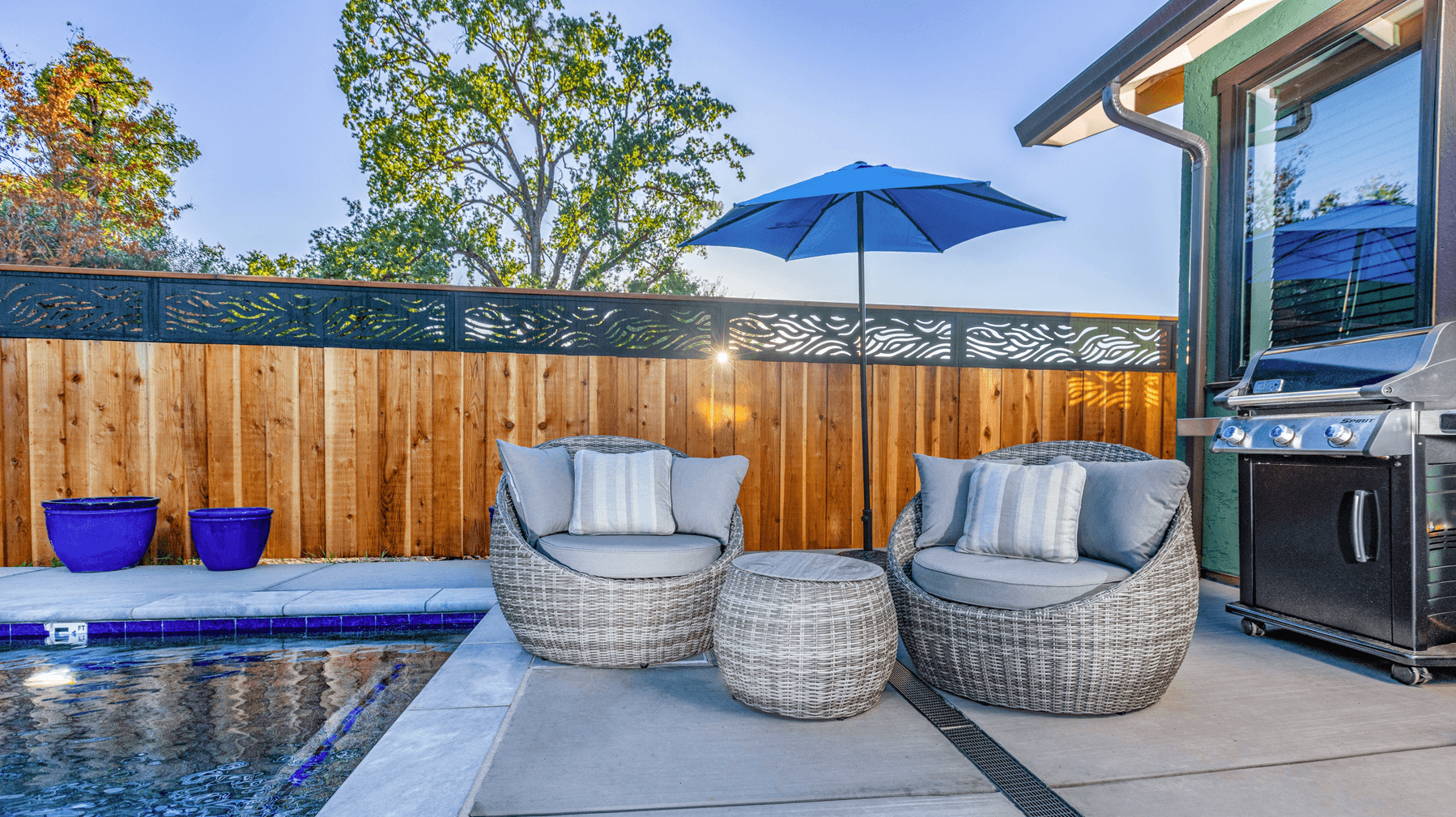 A cozy outdoor seating area with two wicker chairs, a round table, an umbrella, and a grill beside a pool.