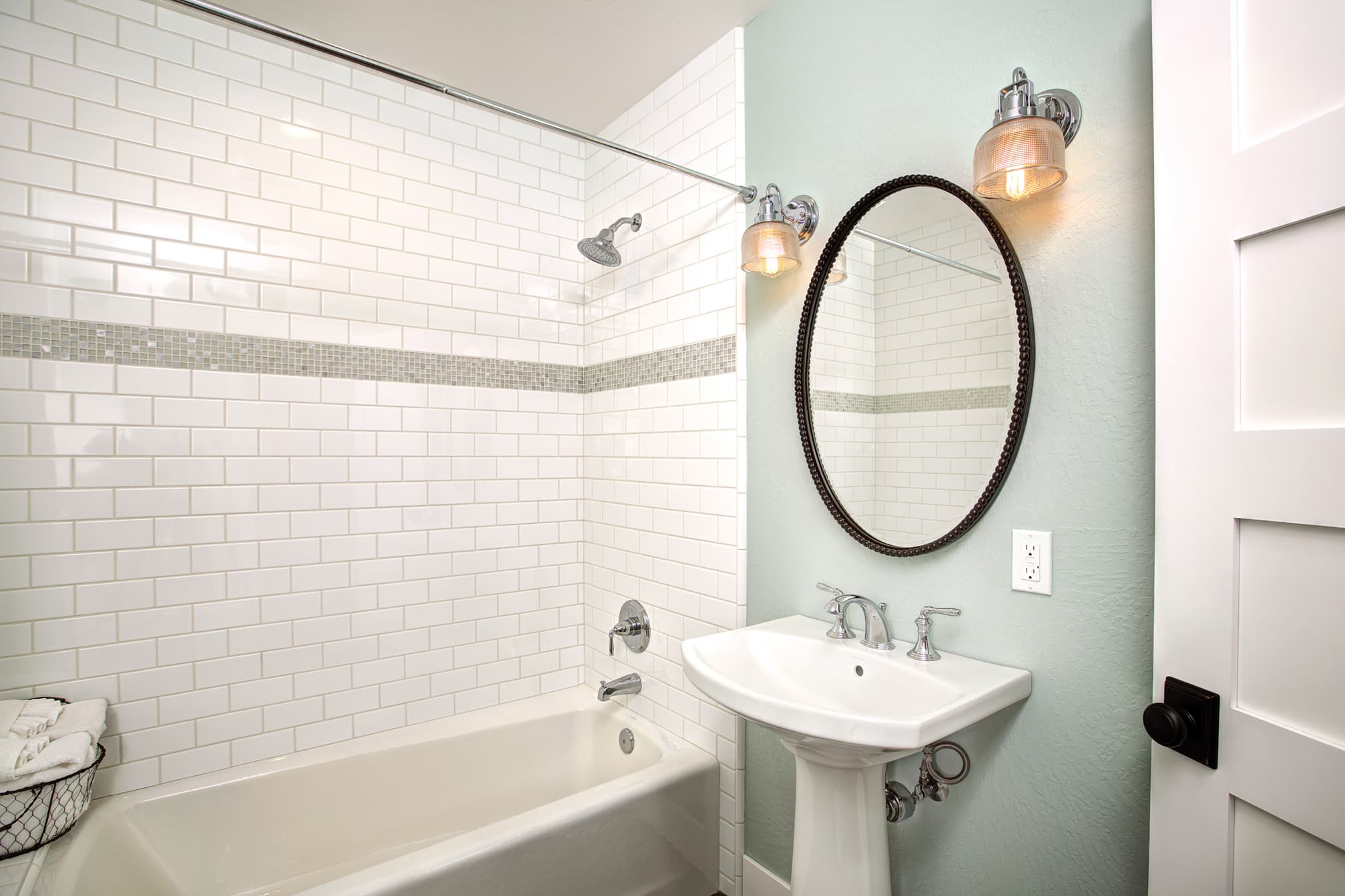 A modern bathroom featuring white subway tiles, a bathtub, a pedestal sink, and a round mirror.