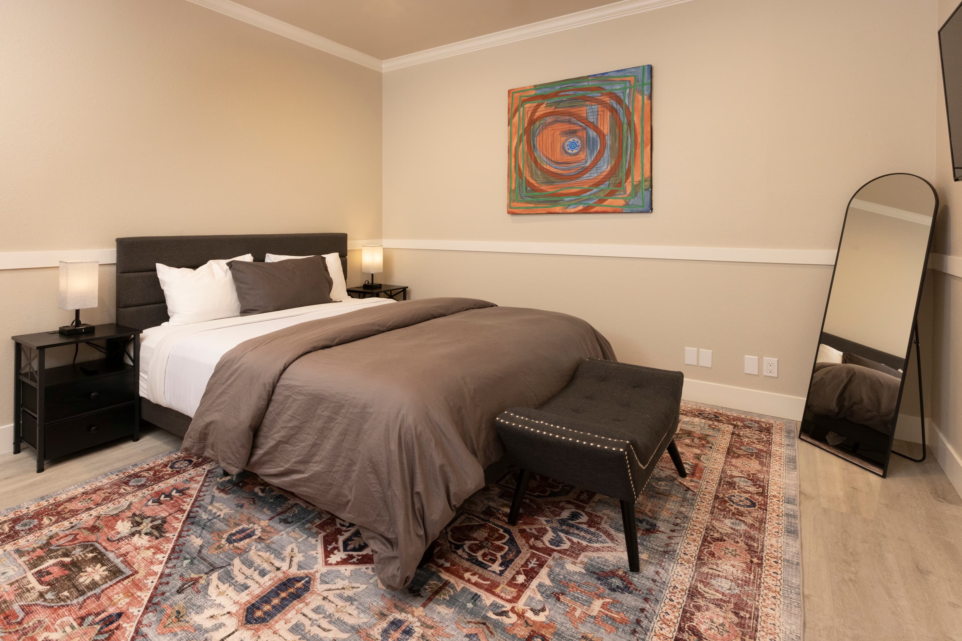 A cozy bedroom featuring a large bed with a gray blanket, decorative artwork, a full-length mirror, and a patterned rug.