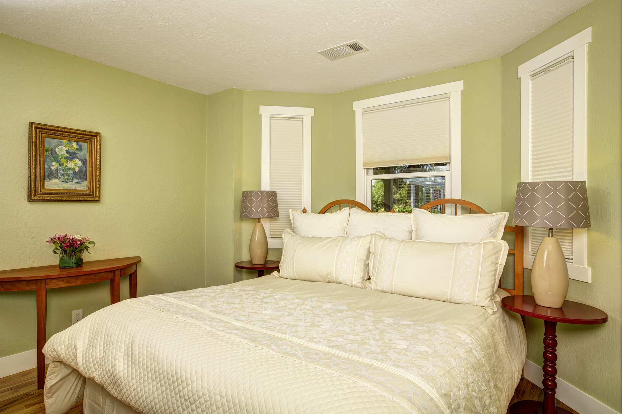 A cozy bedroom featuring a queen-sized bed, two bedside lamps, and a small table with flowers, set against a green wall.