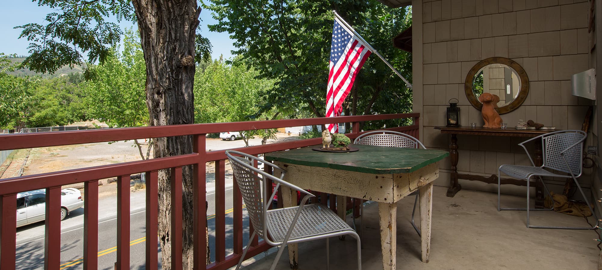 A balcony featuring a green table, chairs, an American flag, and nearby trees.