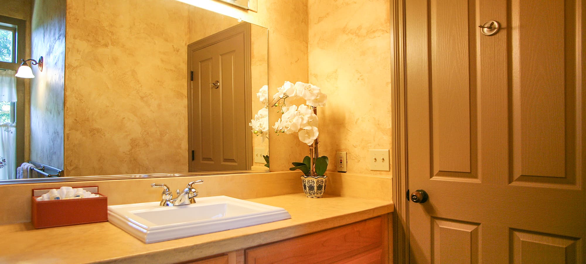 A well-lit bathroom featuring a sink, a vase with white flowers, and a wooden door.