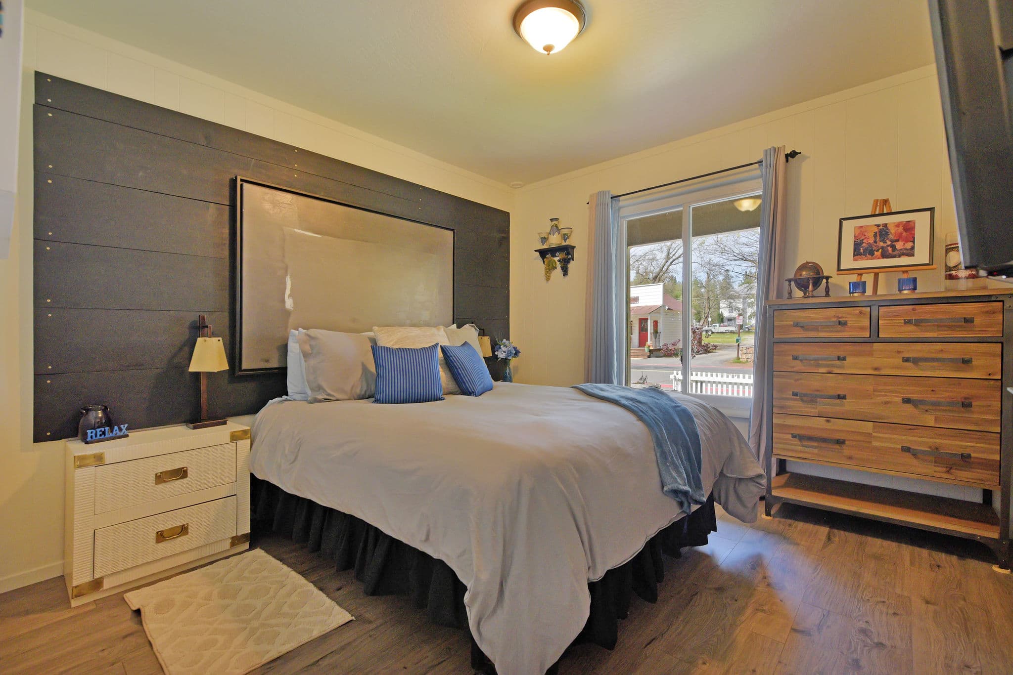 A cozy bedroom featuring a bed with blue pillows, a wooden dresser, and large windows that let in natural light.