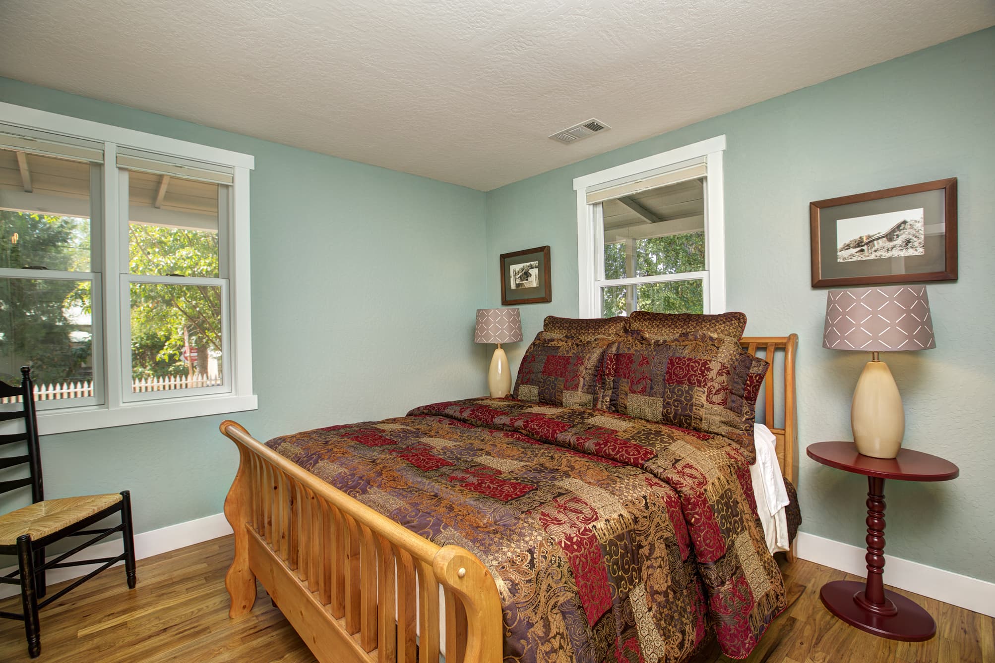 Cozy bedroom featuring a wooden bed with a patterned quilt, two windows, and stylish lamps.