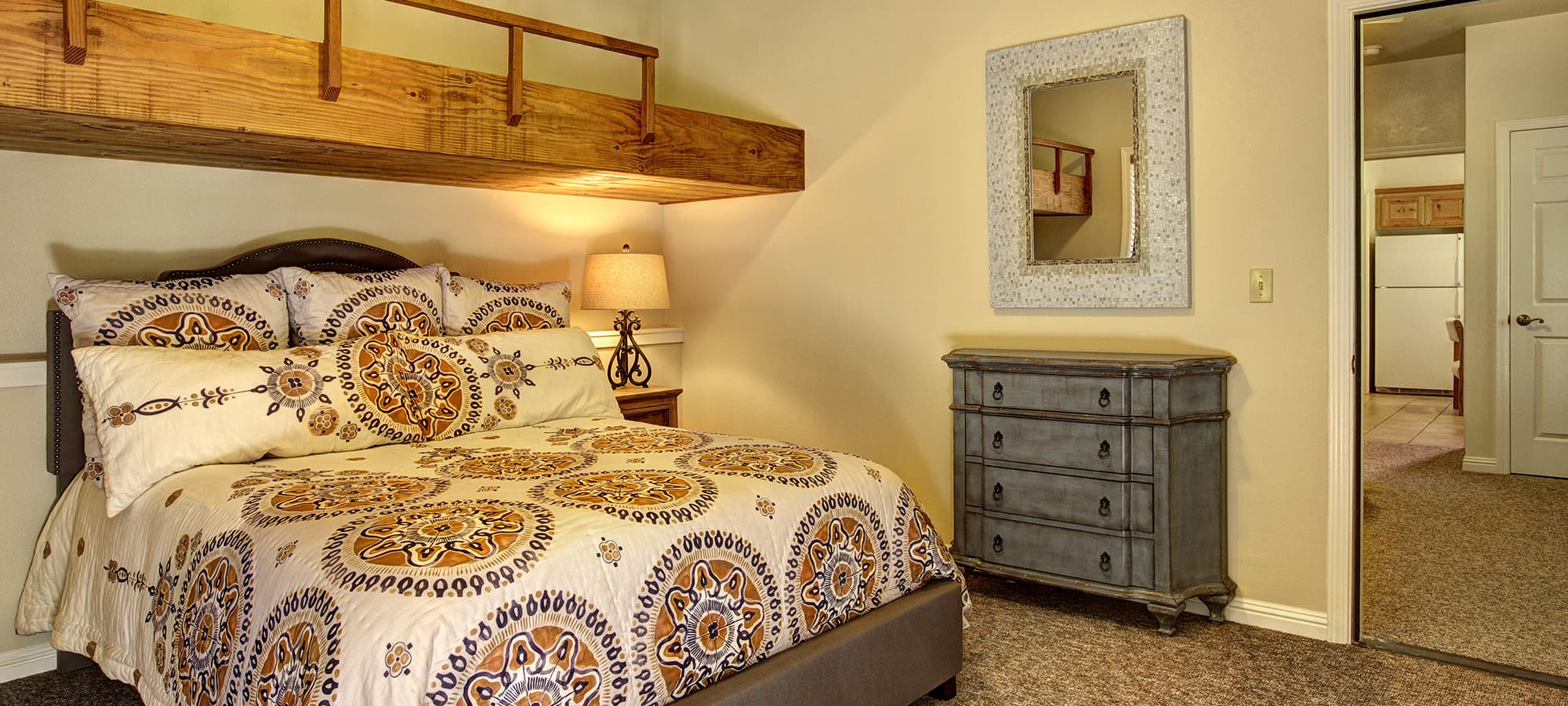 Cozy bedroom featuring a patterned bedspread, lamp, and a mirrored wall decor with an adjacent door leading to another room.