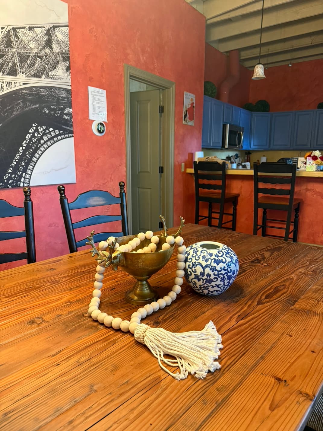 A wooden table with decorative items, including a brass bowl, a beaded garland, and a blue-and-white vase, set against a red wall with kitchen visible in the background.