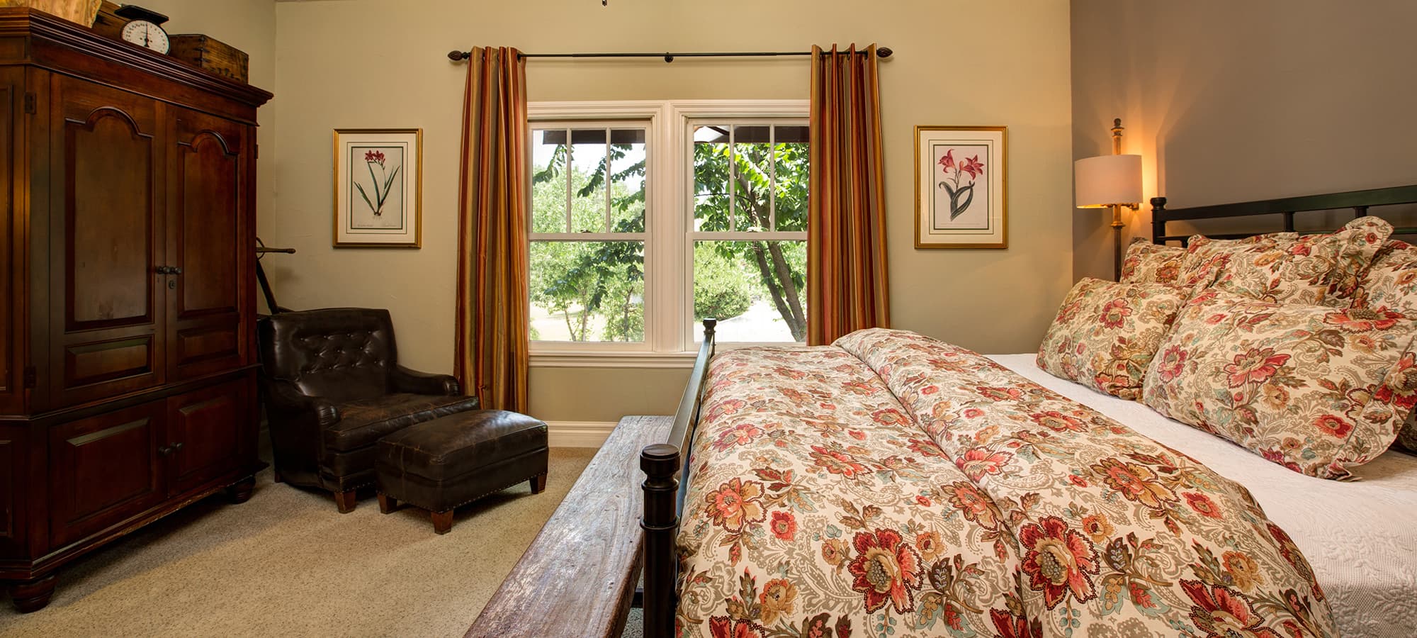 A cozy bedroom featuring a floral-patterned bedspread, a wooden armoire, a leather chair, and a window with drapes.