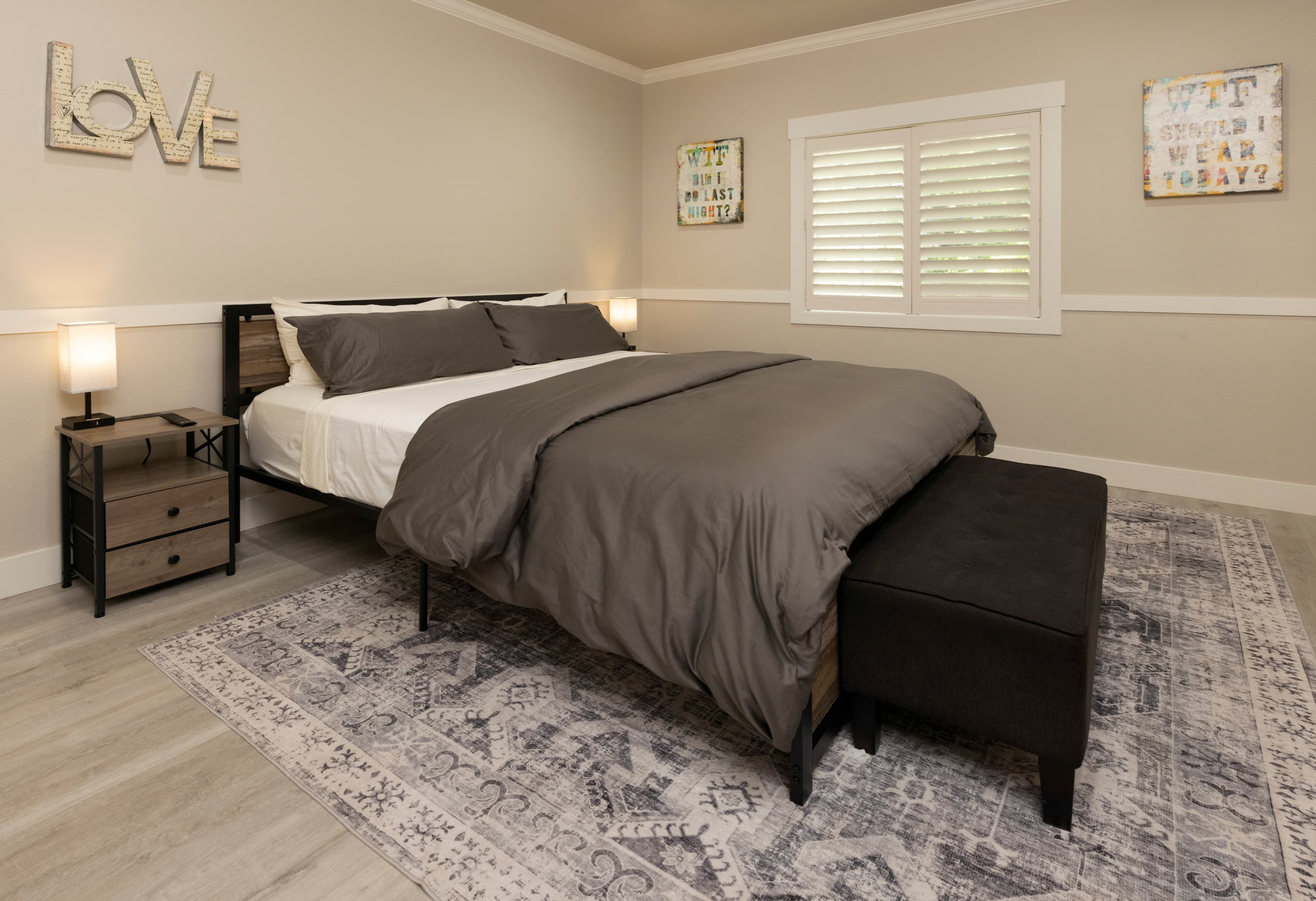 A cozy bedroom featuring a neatly made bed with gray bedding, a dark ottoman at the foot, and decor on the walls.
