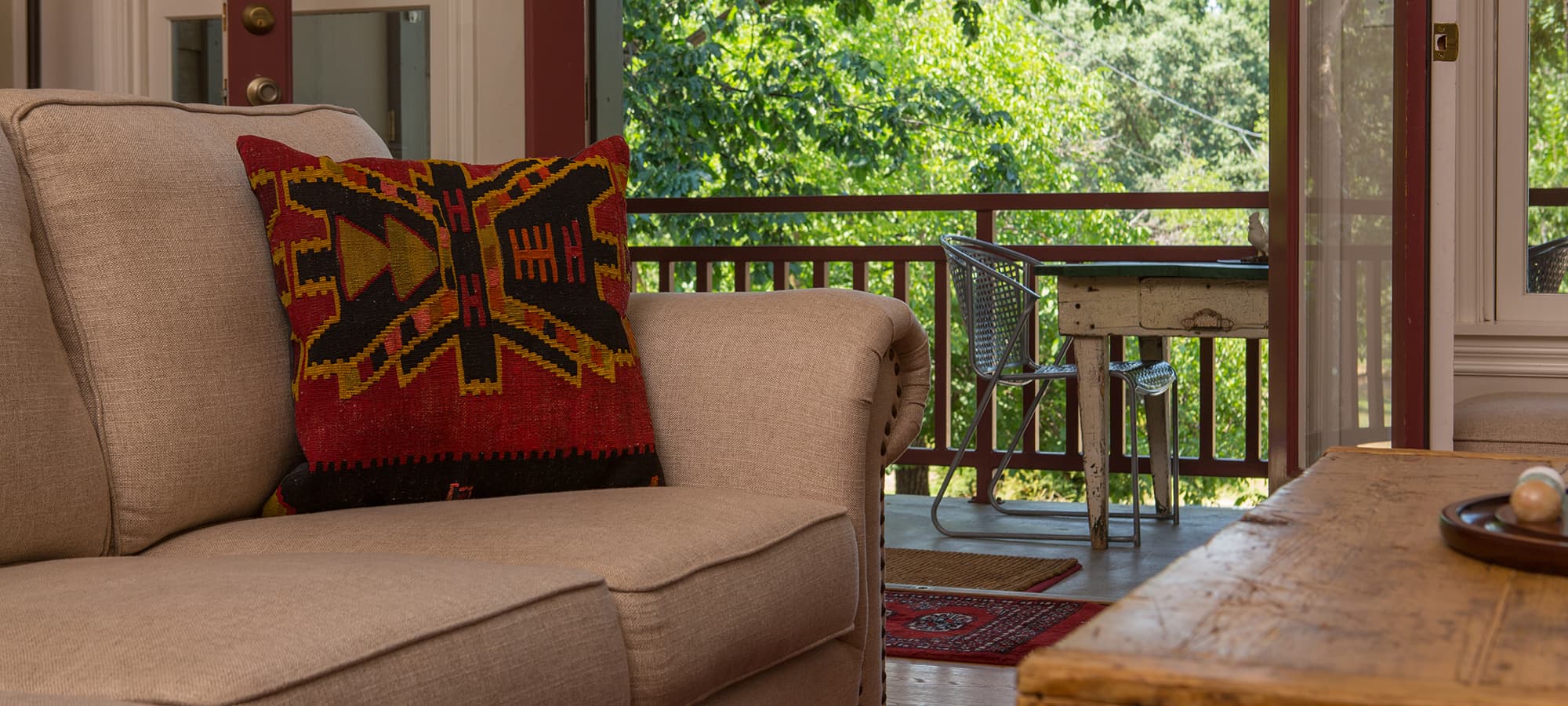 A cozy living space features a sofa with a vibrant patterned pillow and a view of a sunny outdoor deck.