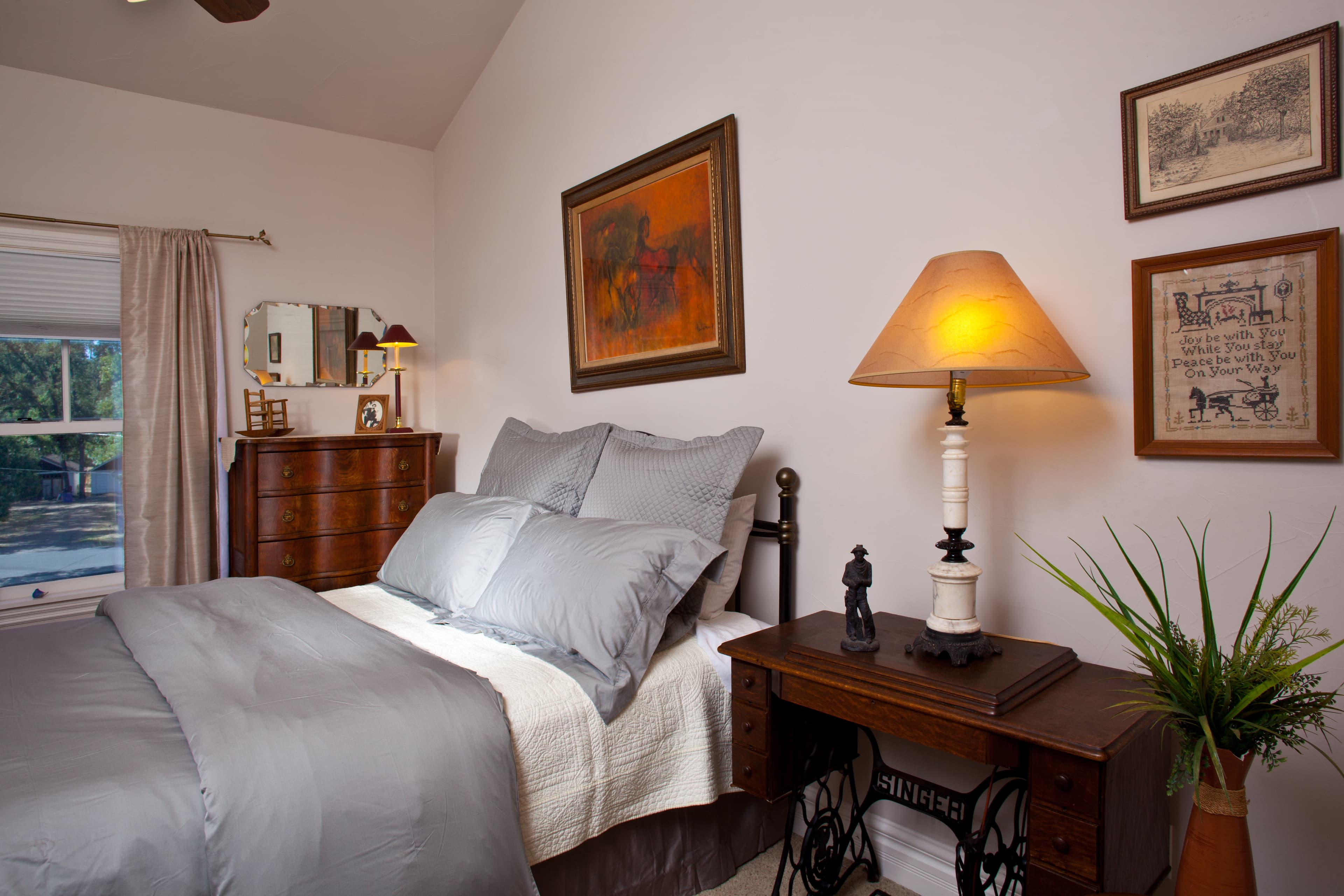 A cozy bedroom featuring a bed with gray linens, wooden furniture, artwork on the walls, and a warm lamp.