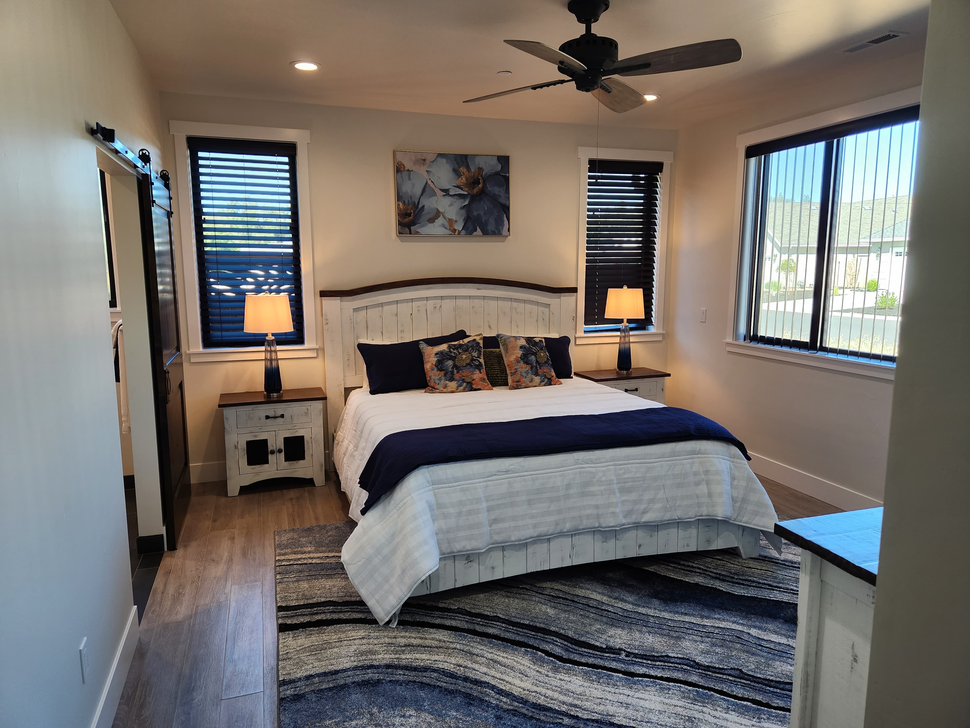 A cozy, well-lit bedroom featuring a queen bed, decorative pillows, nightstands with lamps, and large windows.