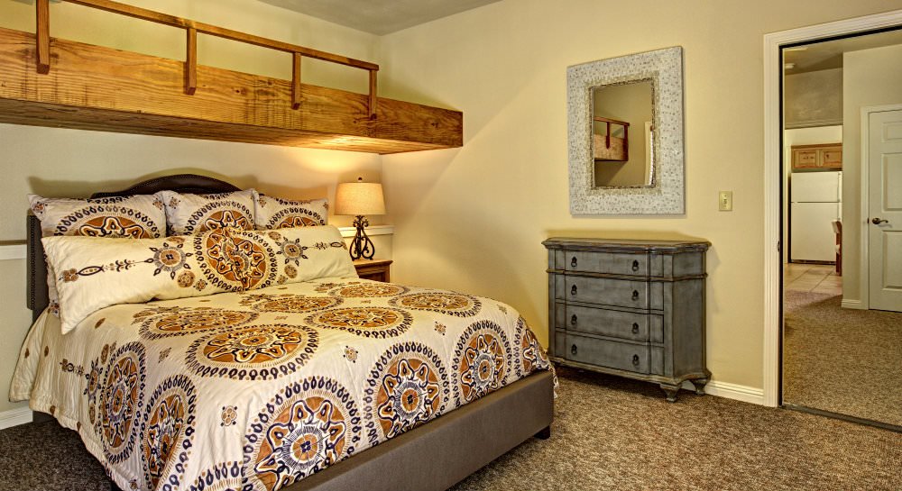 Bedroom with brown carpet, beige walls, large bed with ivory brown and blue comforter, a side bureau and mirror