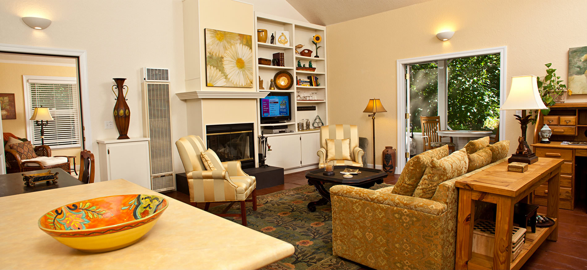 Open area with vaulted ceiling, gold furniture, fireplace, red tile flooring, and kitchen with white cabinets and yellow walls