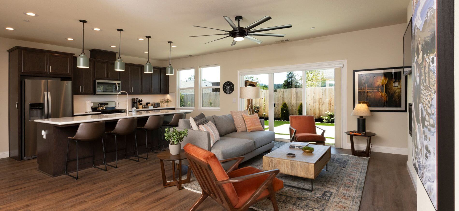 Living room with grey couch & orange chairs facing wall with tv mounted, kitchen with stainless steel appliance and dark cabinets plus island with bar chairs, sliding door to fenced yard.