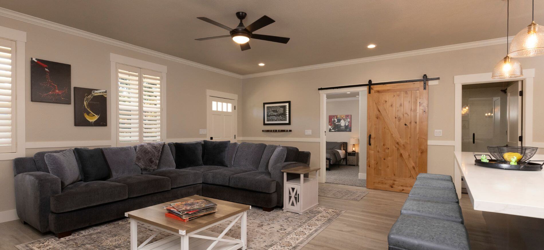 Beige living room with hard floors covered by rugs, dark grey couch in shape of "L", ceiling fan, island with cushions stools, artwork on walls