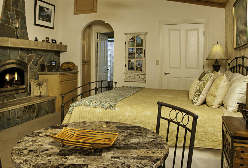 Charming vaulted bedroom with stone fireplace, cream walls, tan carpet and dark metal bed with gold bedspread