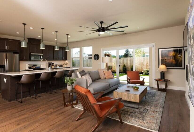 Living room with grey couch & orange chairs facing wall with tv mounted, kitchen with stainless steel appliance and dark cabinets plus island with bar